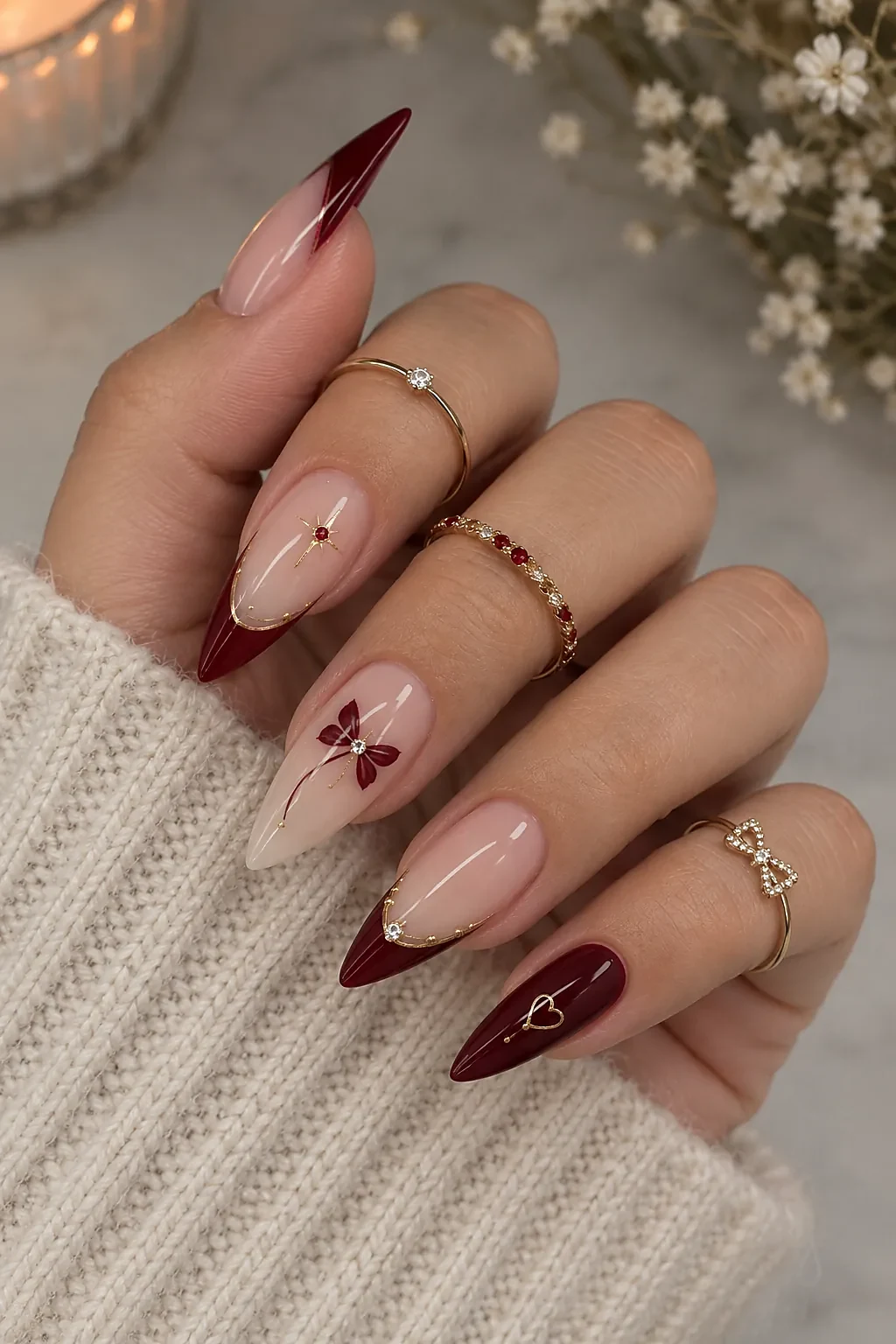 Stiletto nails with nude bases and deep burgundy diagonal French tips, gold outlines, a burgundy flower with a rhinestone, a gold starburst, and a gold heart outline